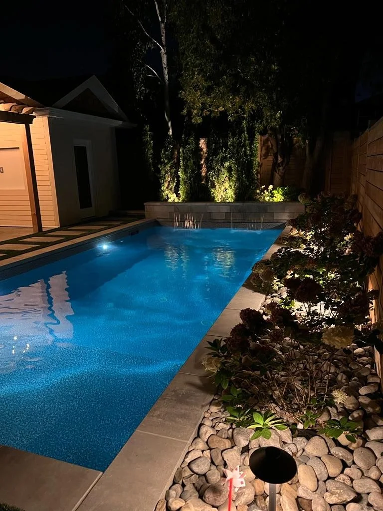 A backyard pool illuminated at night with surrounding landscaping including bushes, rocks, and trees, and a wooden fence. Lights are directed at the plants and trees.