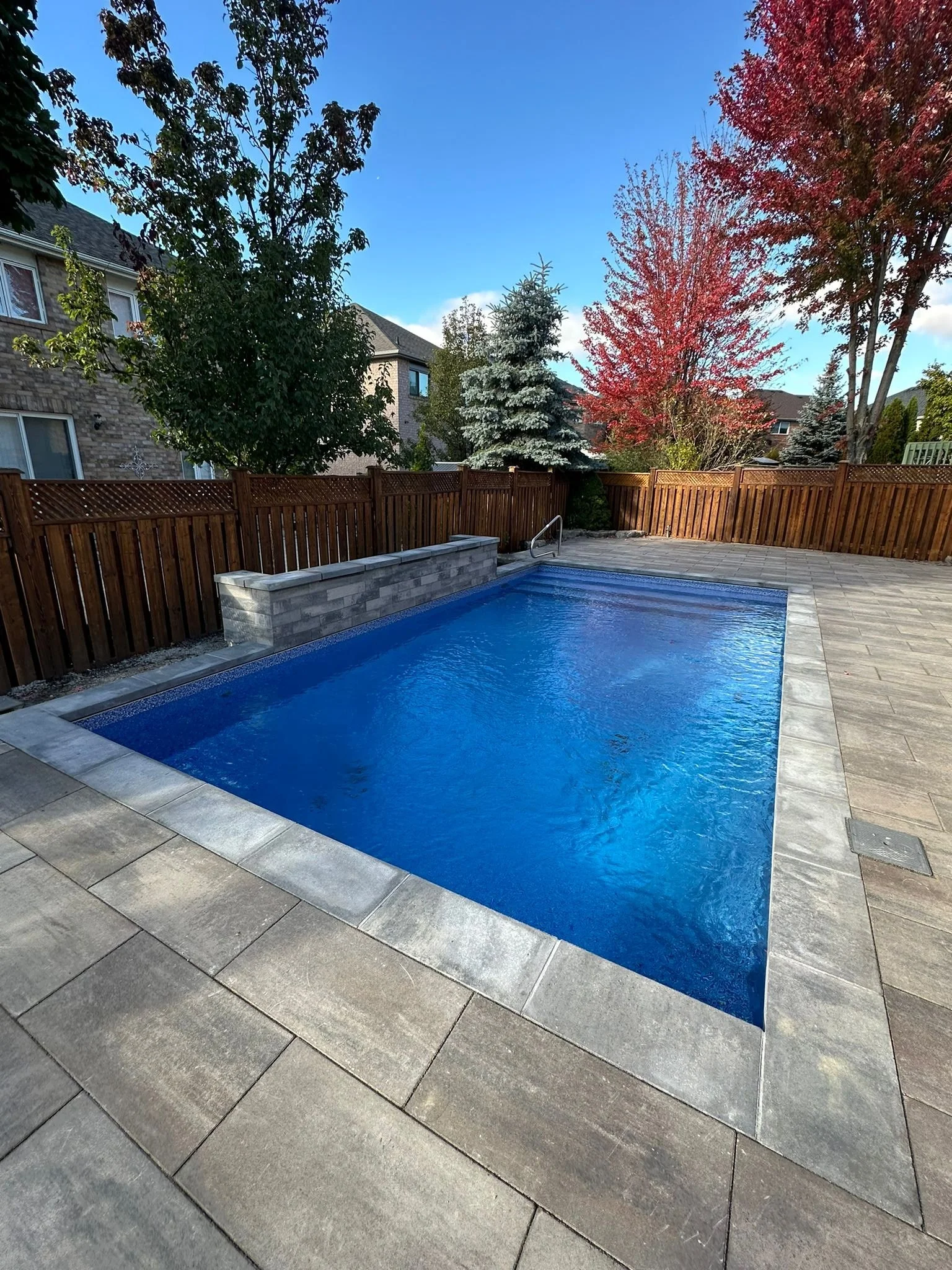 A backyard swimming pool with blue water, surrounded by stone pavers, with a wooden fence, trees, and a clear sky in the background.