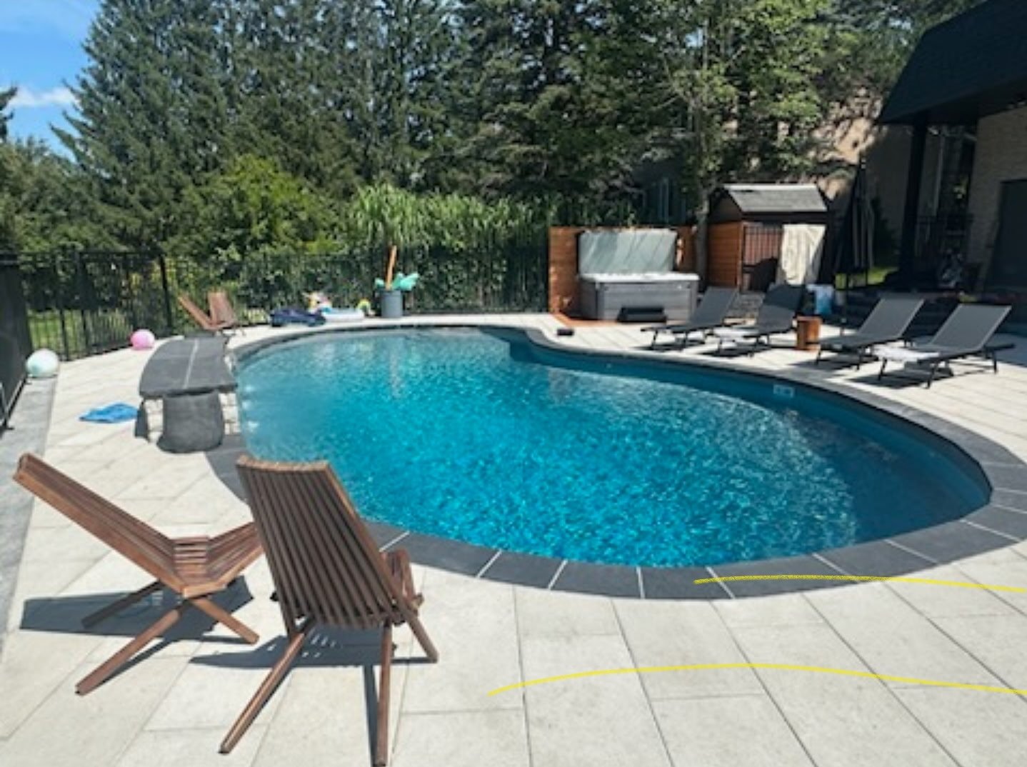 A backyard with a curvy in-ground swimming pool, surrounded by a paved deck with lounge chairs, a shed, trees, and a black metal fence, on a sunny day.