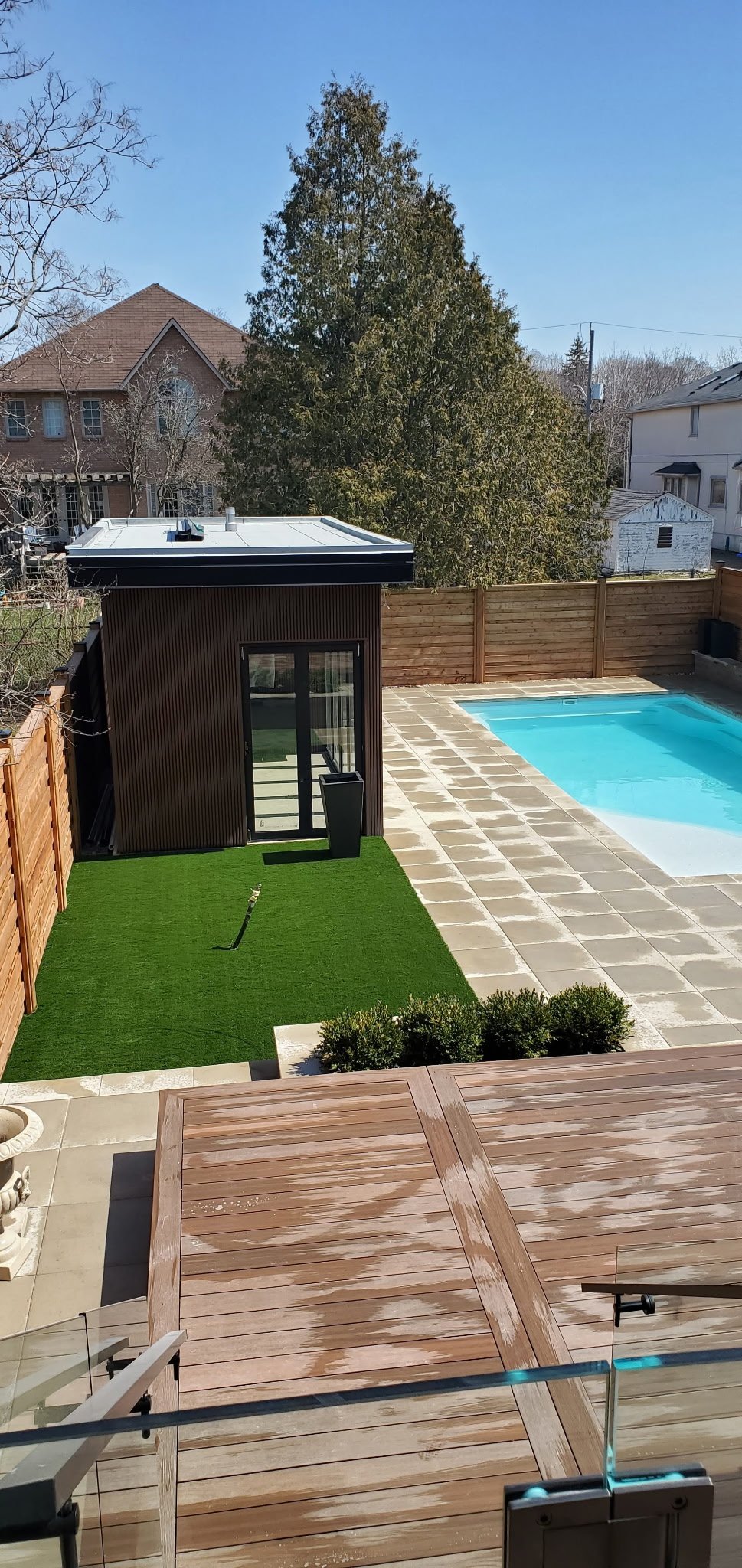 A backyard with a swimming pool, a small black shed, artificial grass, a wooden deck, and a tall tree in the background under a clear blue sky.