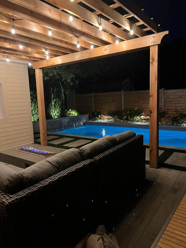 Nighttime backyard scene featuring a lit string light pergola over a sitting area with a cushioned outdoor sofa. In the background, there's a blue illuminated swimming pool with a wooden fence and some plants along the fence line.