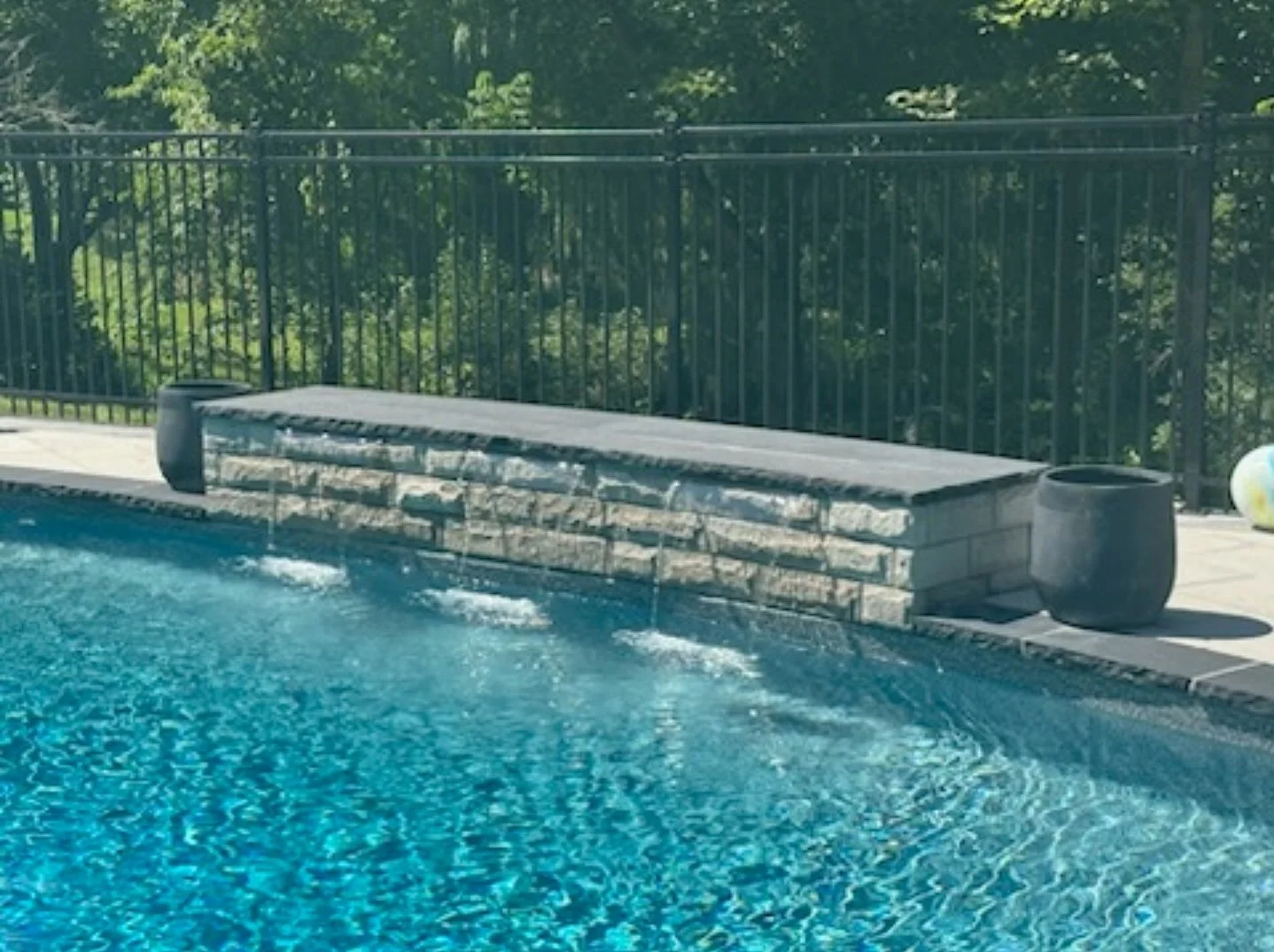 View of a backyard swimming pool with a water fountain feature, surrounded by a black metal fence, potted plants at the edge, and lush green trees in the background.