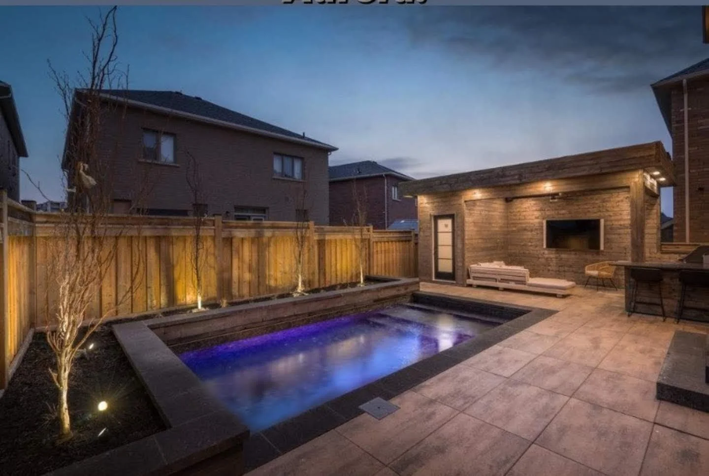 Backyard patio with a lit pool, wooden fencing, a small brick shed with outdoor seating, and houses in the background during dusk.