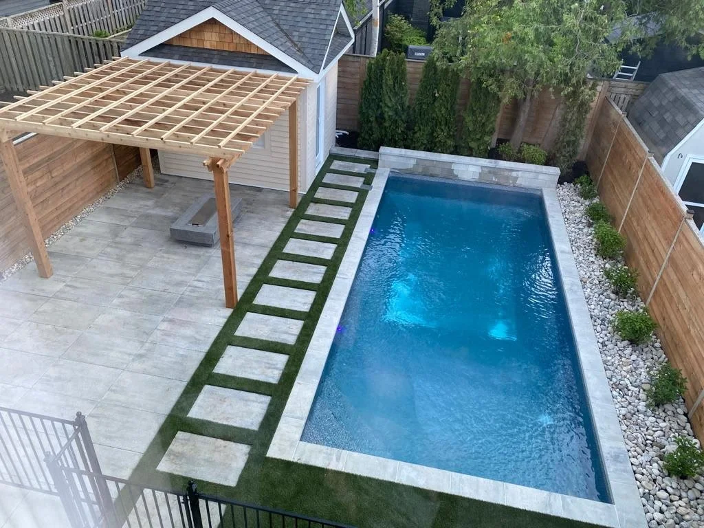 Backyard with a rectangular swimming pool, a wooden pergola, a small shed, artificial grass pathway, and surrounding fence with greenery.