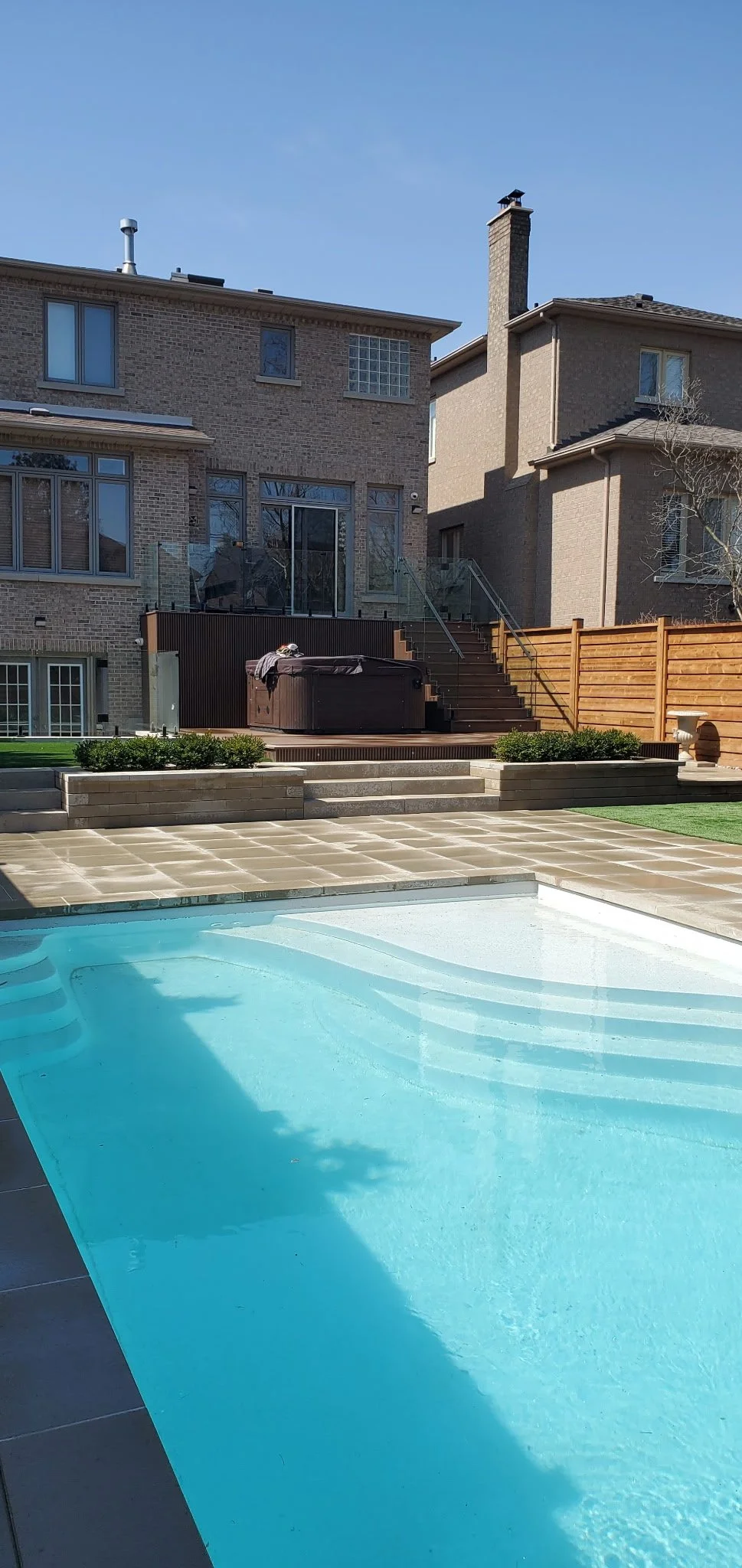 Backyard with swimming pool, patio, small hot tub, raised deck with stairs, brick houses, wooden fence, and some greenery under a clear blue sky.