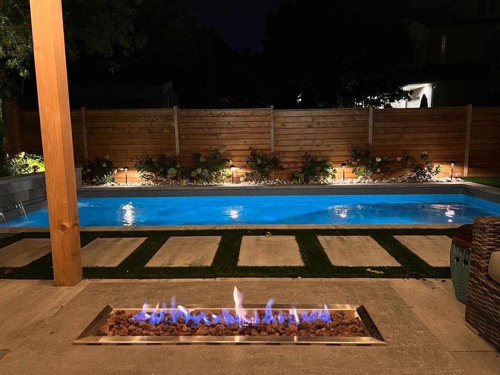 Nighttime backyard with a lit rectangular swimming pool, a wooden privacy fence, a fire pit with blue flames, and a patio with chairs.