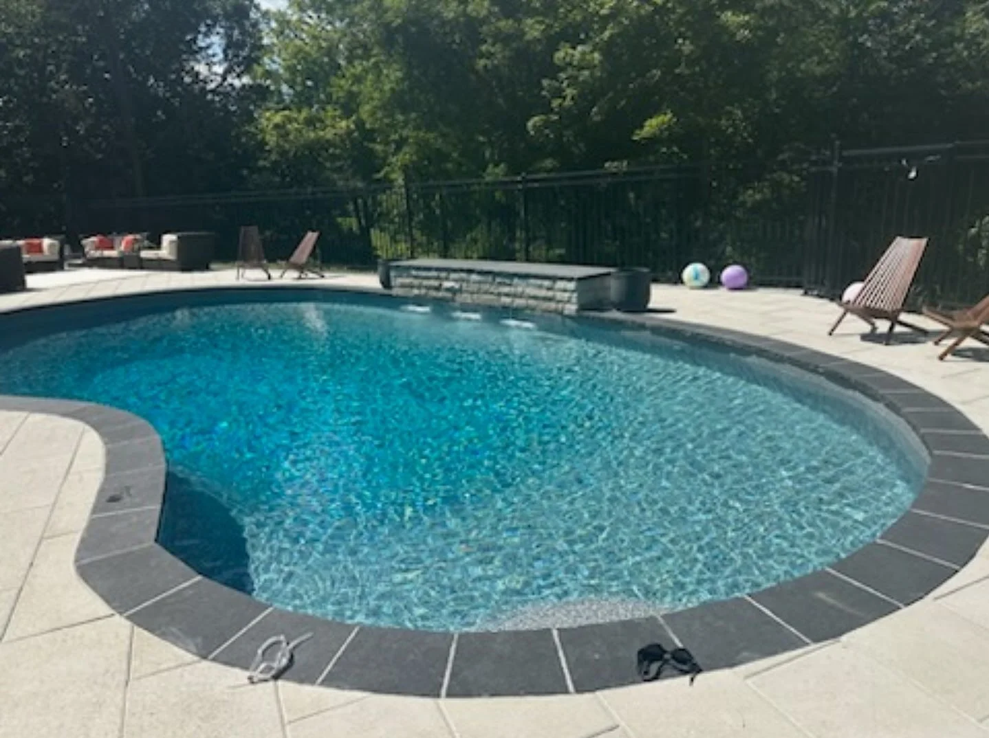 Oval-shaped swimming pool with blue water, surrounded by a concrete deck and black border. Poolside chairs and some colorful balls are on the deck, with trees and a black metal fence in the background.