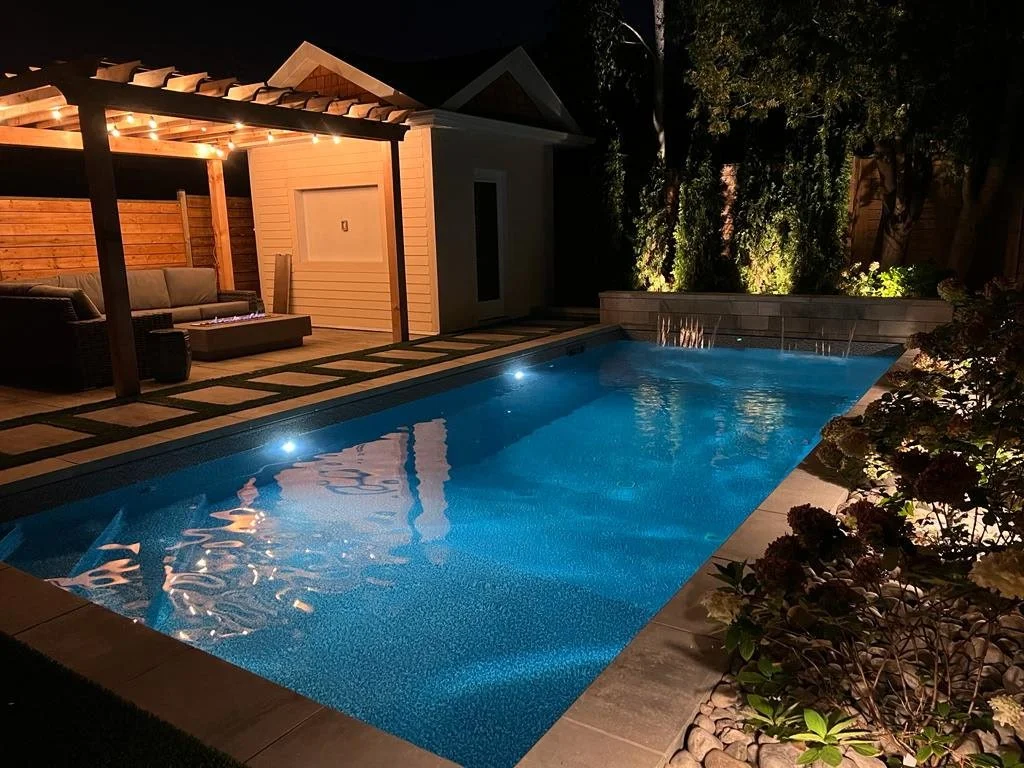 Nighttime view of a backyard swimming pool illuminated with underwater lights, surrounded by plants and trees. There is a covered patio with outdoor seating and string lights.