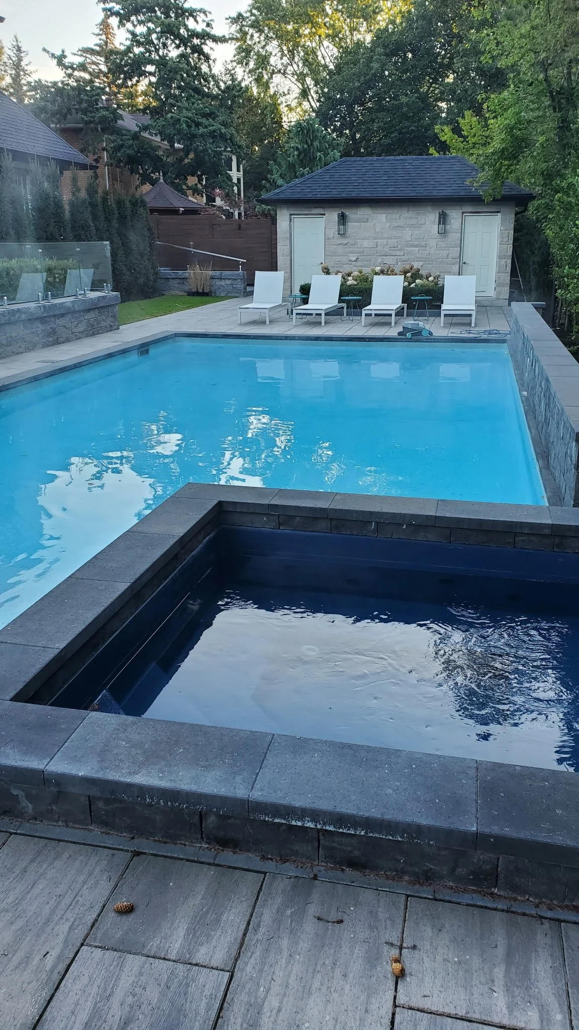 A backyard swimming pool with a hot tub in the foreground, surrounded by stone pavers, with a small building and four lounge chairs in the background, surrounded by trees.