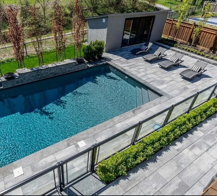 A backyard with a rectangular swimming pool, lounge chairs on a tiled deck, a small building or shed, and a glass fence with green bushes in front.