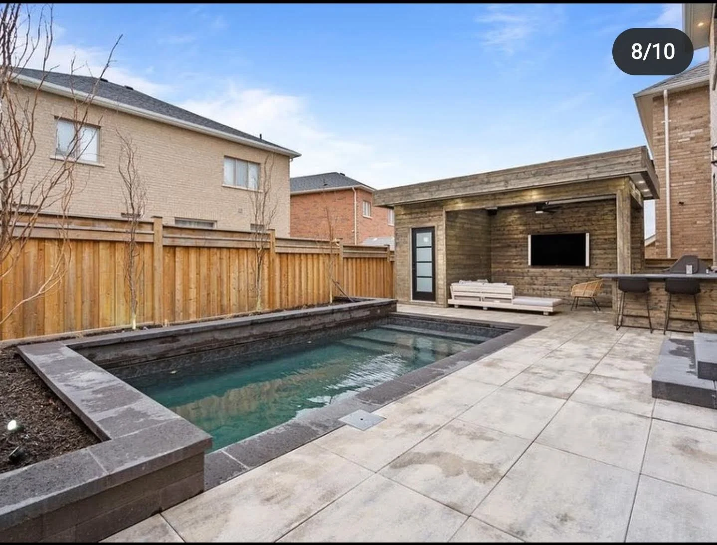 Residential backyard featuring a small rectangular swimming pool with stone border, wooden privacy fence, a wooden pool house with a mounted flat-screen TV, a white outdoor sofa, two barstools at a counter, and a tree without leaves.