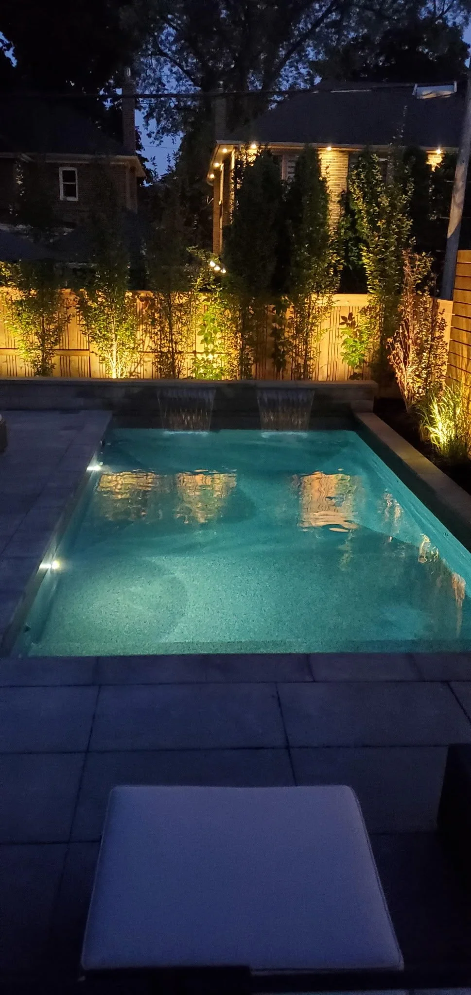Nighttime view of a backyard pool with illuminated water features, surrounded by a wooden privacy fence and garden plants, with a house illuminated in the background.
