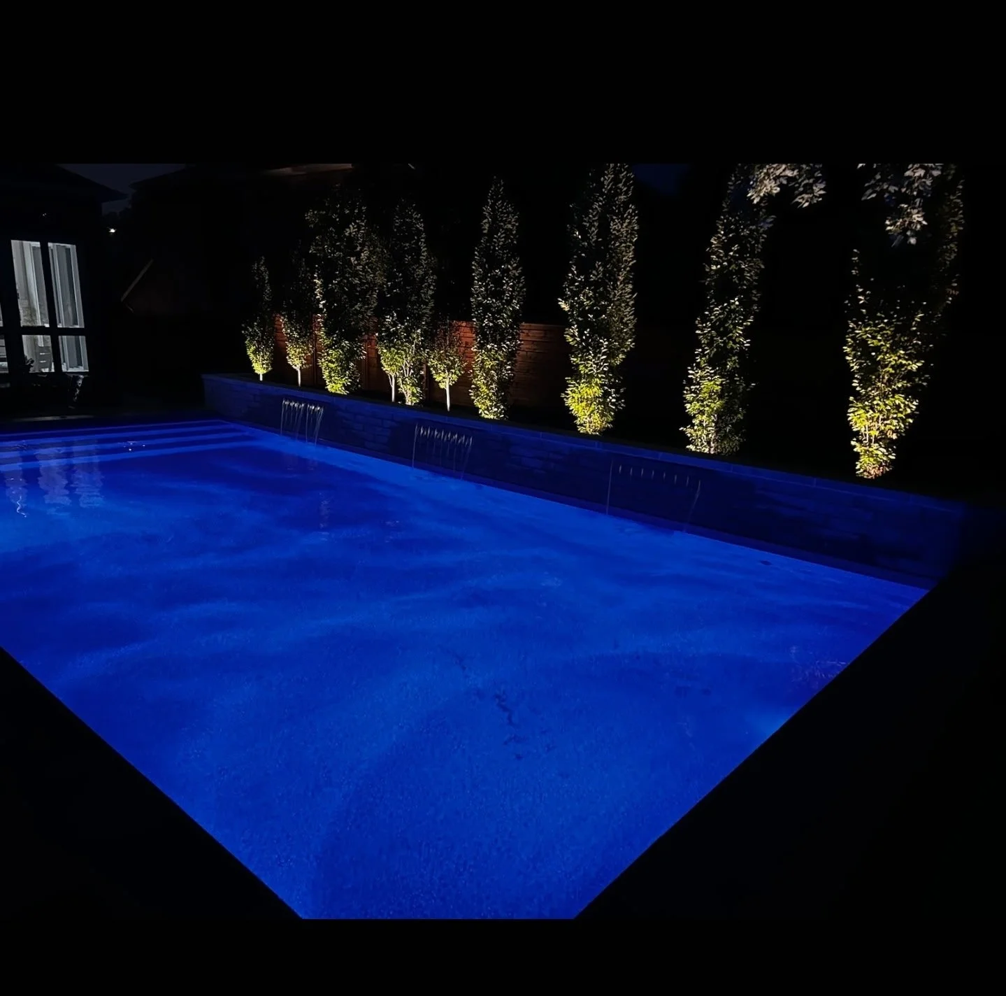 Night view of a lit-up swimming pool with blue water, water features, and a row of illuminated trees in the background.
