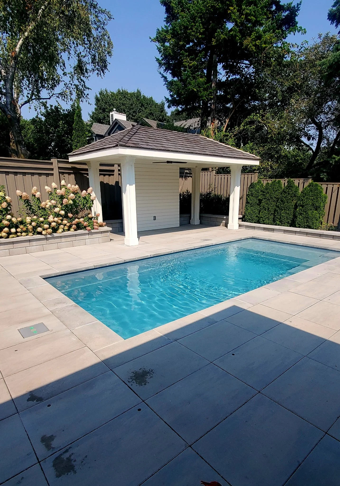 Beautiful backyard with a swimming pool, tiled patio, small white pool house, flowers, and tall trees in the background.