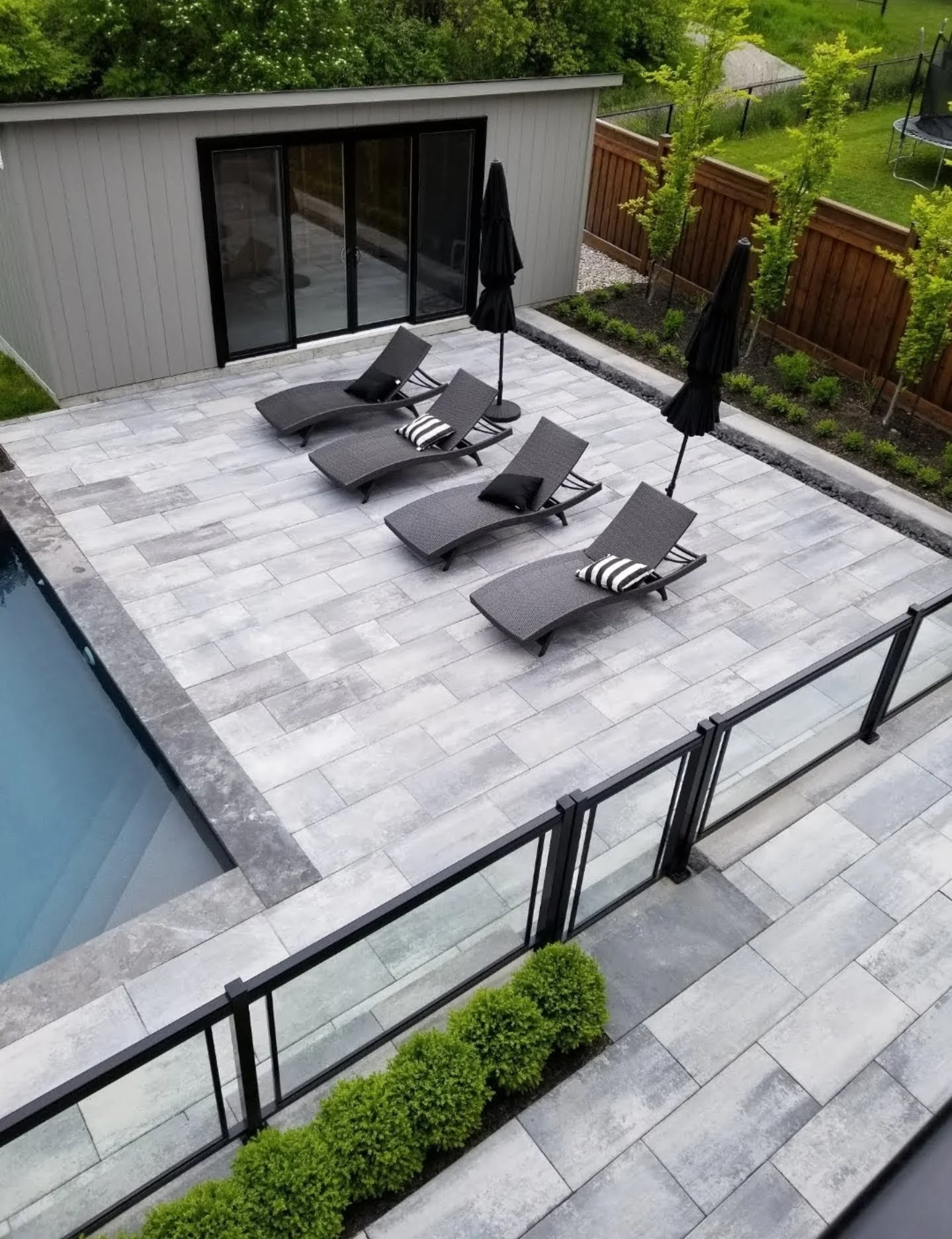 A modern backyard patio with four black lounge chairs, three black umbrellas, and two small striped pillows. The area is enclosed by a glass fence, with a small garden along the fence line and adjacent to a modern gray building with sliding glass doors.