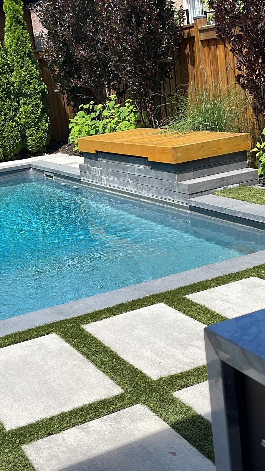 A backyard swimming pool with clear blue water, surrounded by gray stone tiles and a concrete patio with grass accents. There is a raised wooden deck with some plants behind it, and a wooden fence in the background with tall bushes and trees.