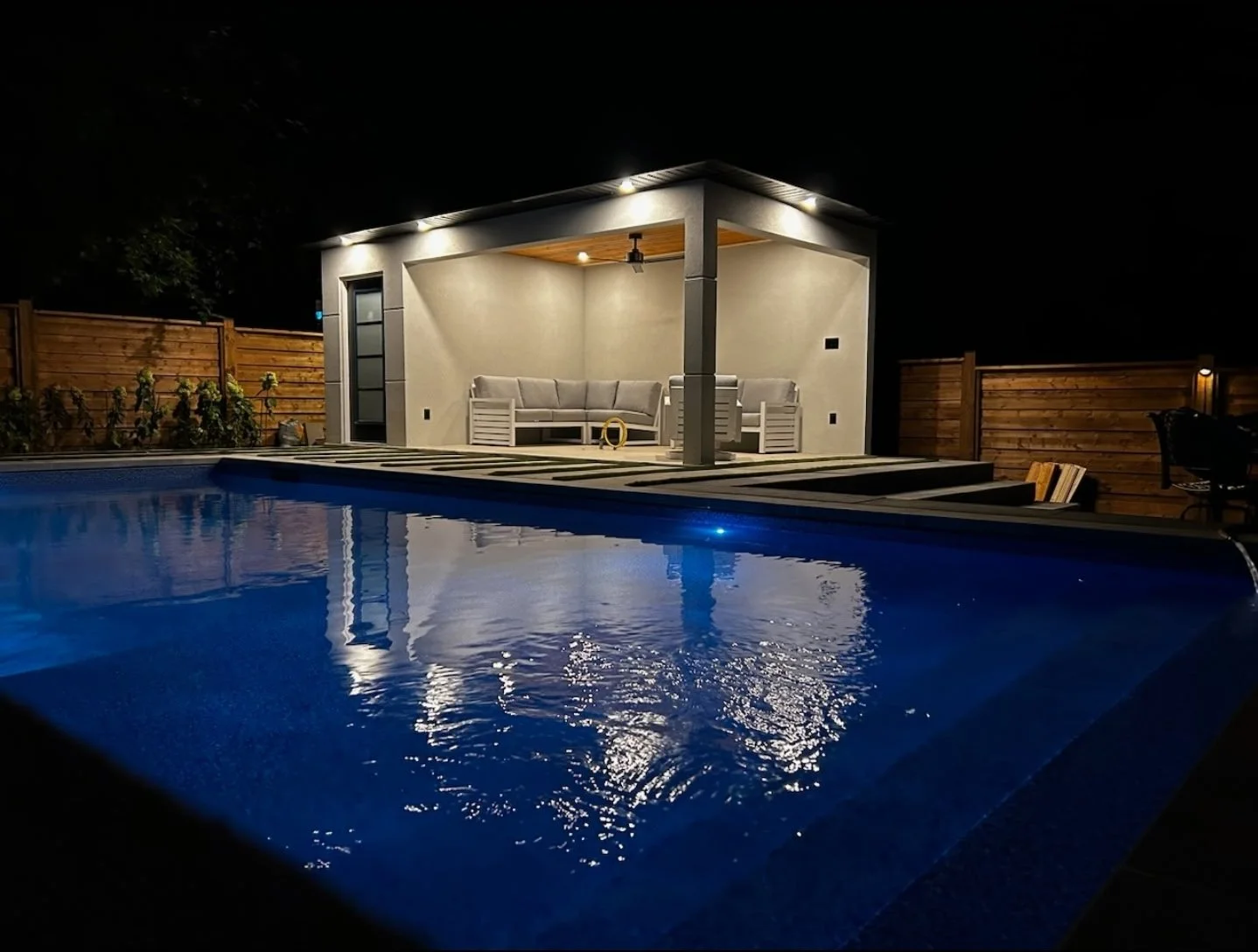 Night view of a backyard with a lit swimming pool in the foreground and a small covered patio with beige seating in the background, surrounded by a wooden fence.