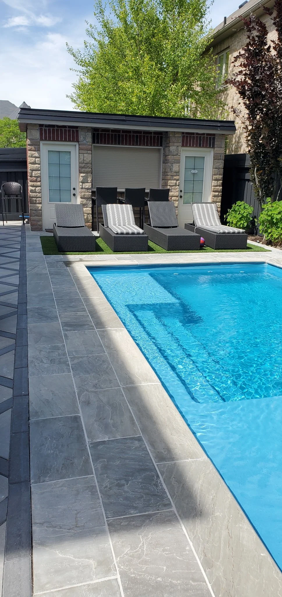 A backyard pool with clear blue water and tiled steps, surrounded by gray stone tiles. In the background, there are four lounge chairs with striped cushions, a small patch of artificial grass, and a stone building with two glass-paneled doors and a window. There is also a patio table with six black chairs, partially visible, and some greenery including a large tree and bushes.