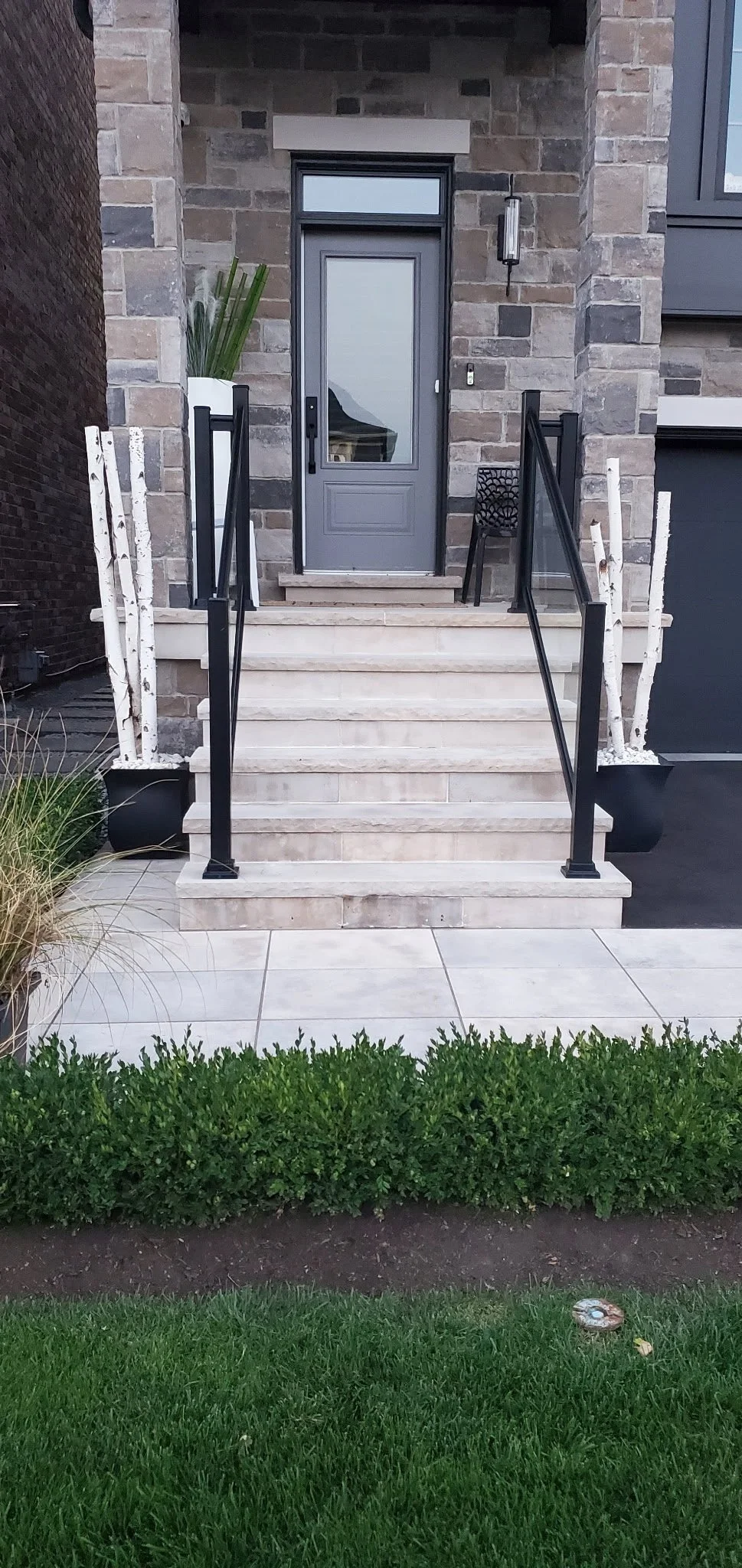 Front porch with stairs leading to a door, plants on either side, and a small bench on the right, with a well-maintained lawn in the foreground.
