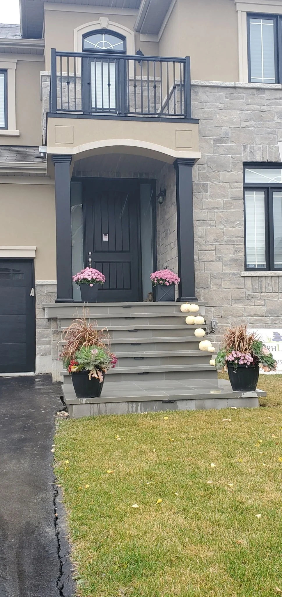 Front porch of a modern house with a black door, gray stairs, black columns, pink potted flowers, and white pumpkins. The house has stone and beige siding, and a small upstairs balcony.
