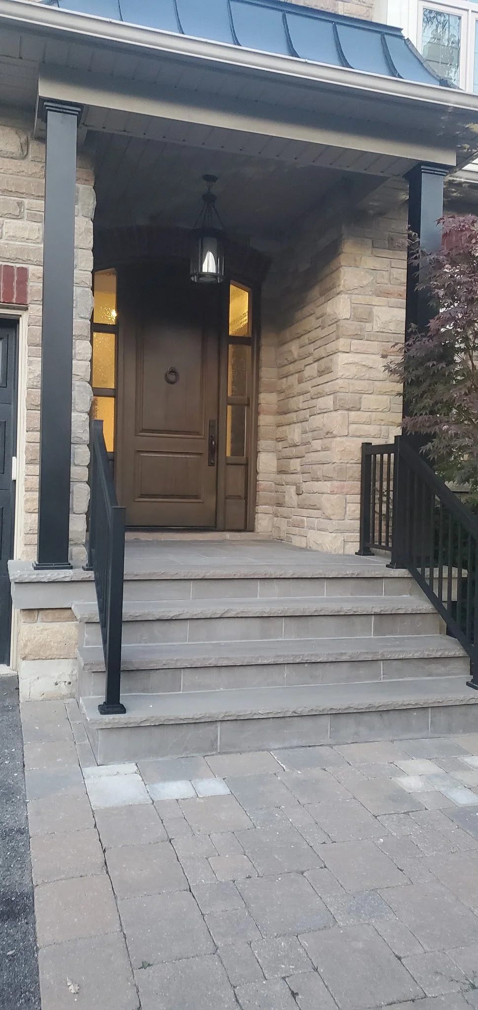 Front porch with stone steps, black railings, and a wooden front door with glass panels, topped by a lantern-style light fixture, and a stone and brick exterior wall.