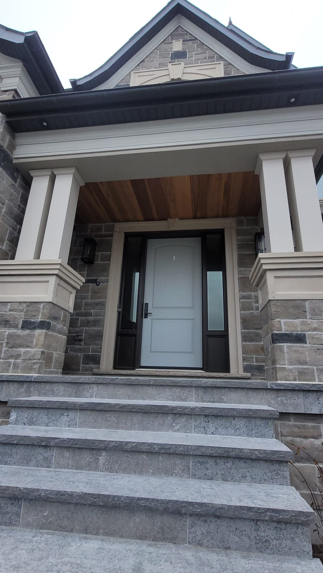 Front entrance of a house with stone steps leading to a white front door, flanked by glass side panels, and a porch with white columns and a wood ceiling.