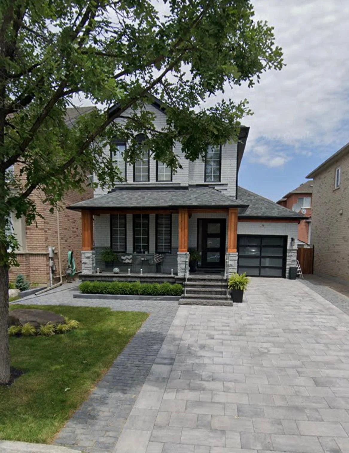 Front view of a modern two-story house with a gray exterior, black trim, and a gray garage door, featuring a front porch with wooden support beams, steps leading up to it, and a landscaped front yard with a grassy area, small plants, and paved driveway.