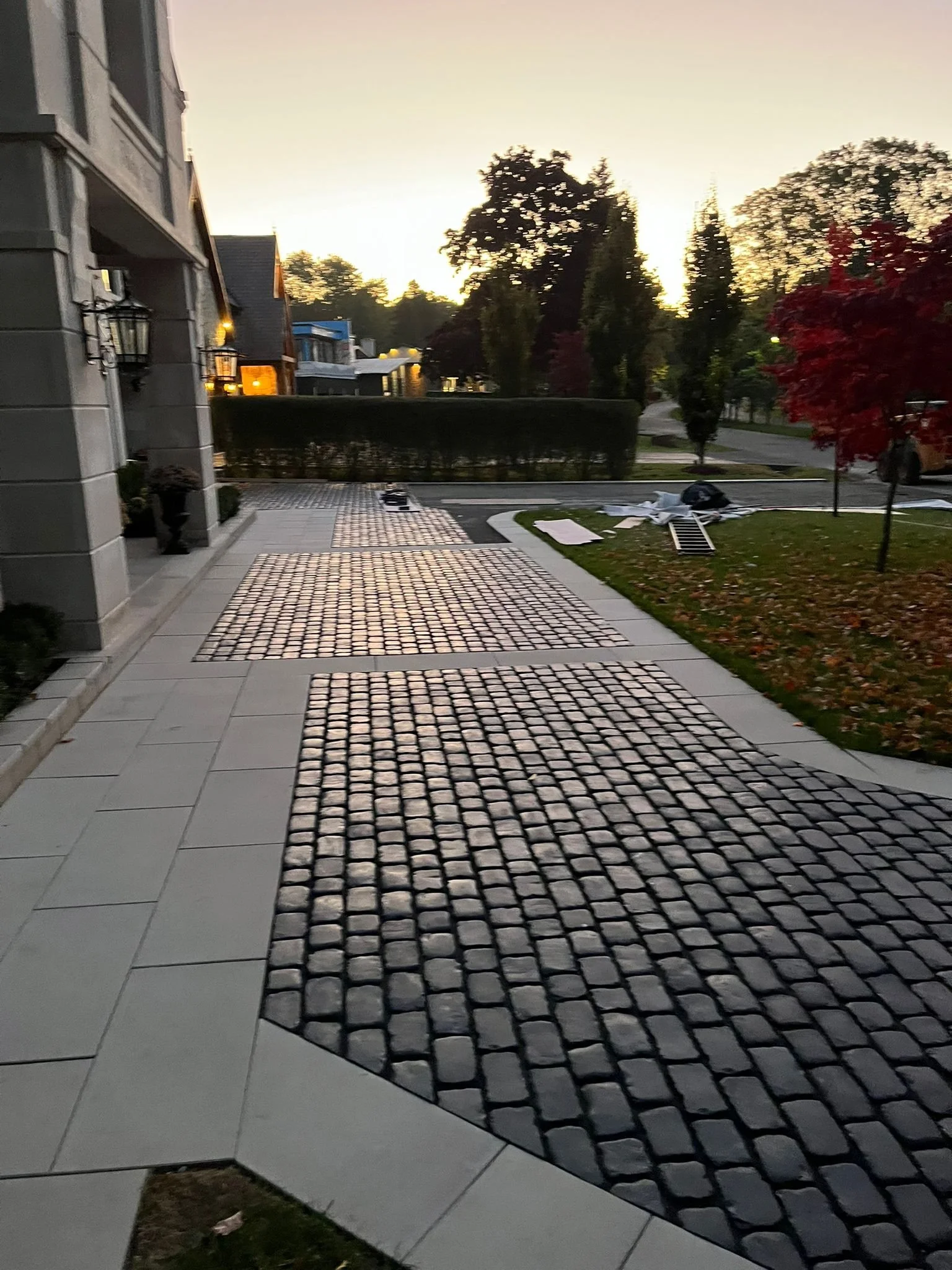 Residential driveway with a combination of light-colored paving stones and dark cobblestones, leading to a house with exterior lighting at dusk, and trees with fall foliage in the background.