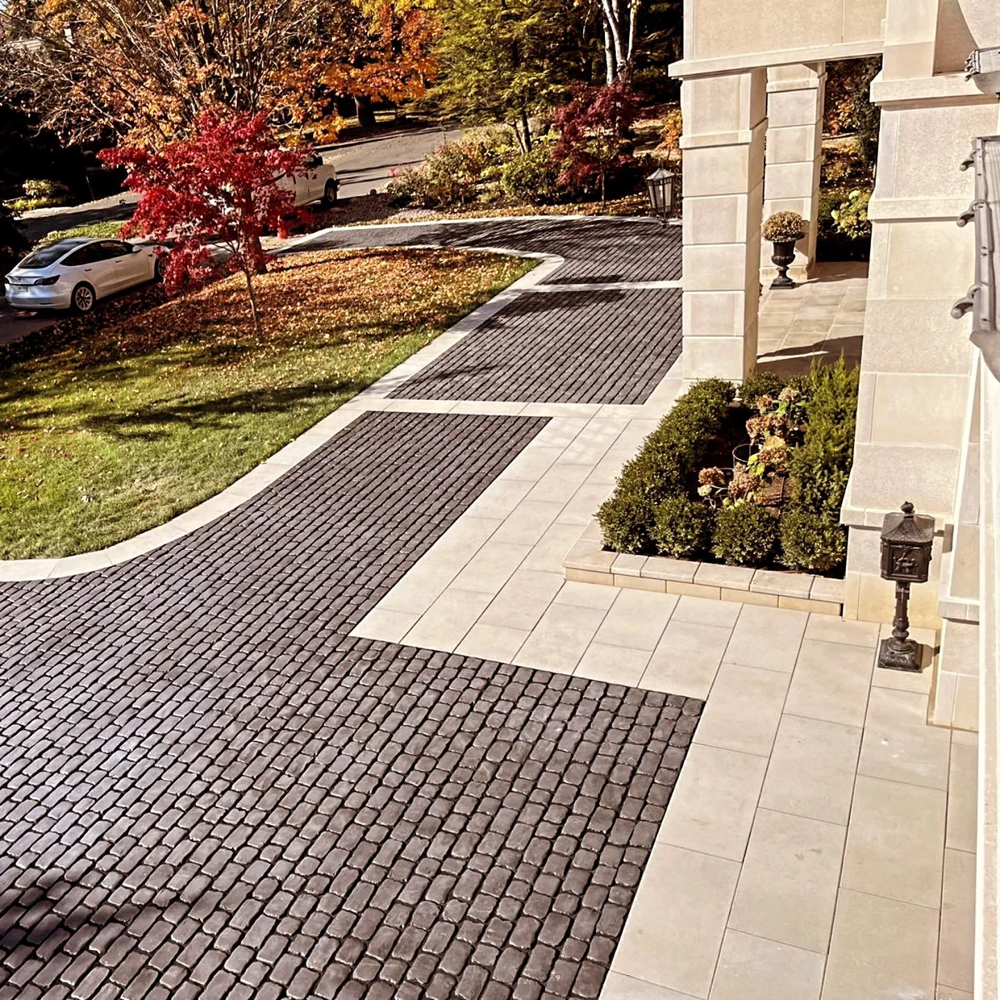 A residential driveway with a cobblestone pattern, bordered by a tiled walkway and landscaped garden with bushes and potted plants, and a street with cars and colorful trees in the background.
