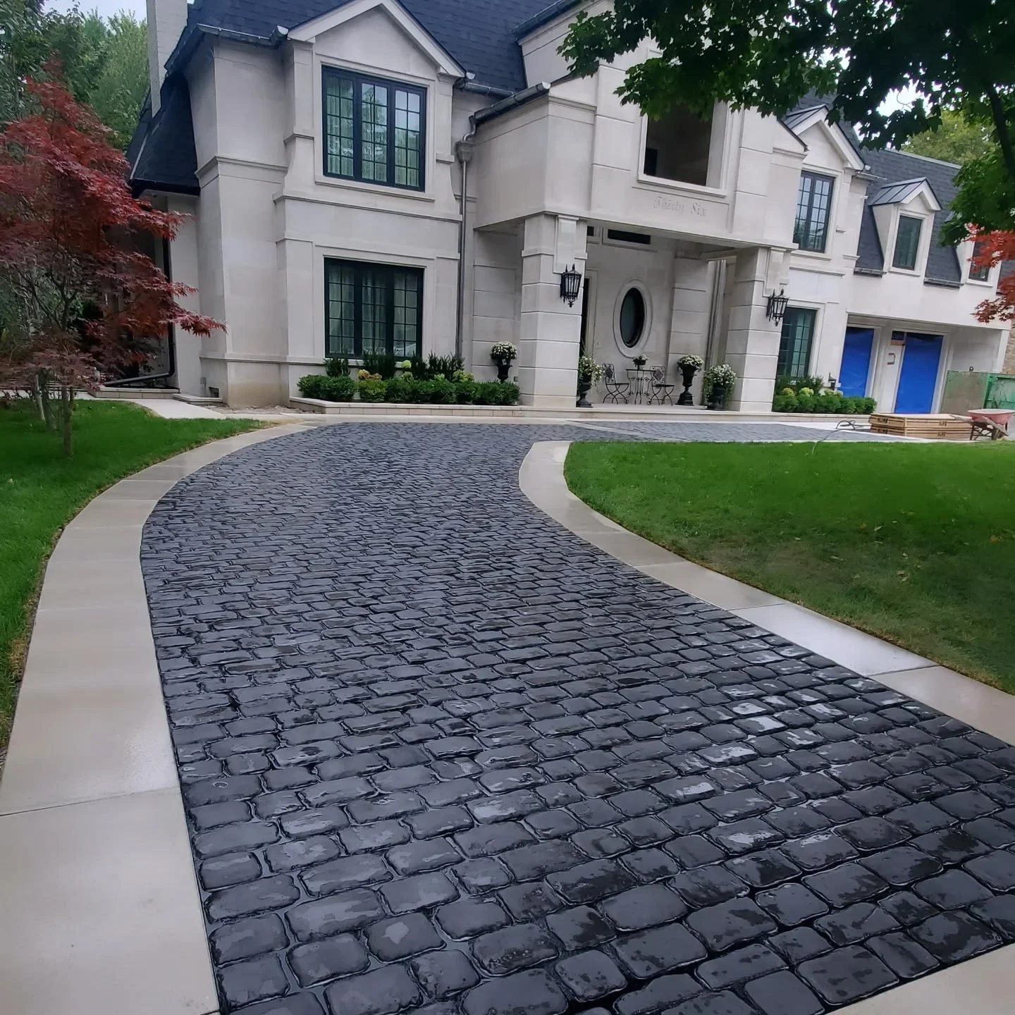 A curved driveway made of dark cobblestone leads to a large, elegant white house with black window frames, a central front door, and outdoor decorations including potted plants and lanterns.