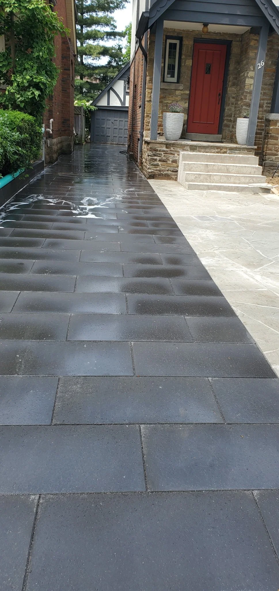 A wet outdoor walkway with dark gray tiles on the left side and lighter gray tiles on the right, leading to a house with a red door and two potted plants on steps.
