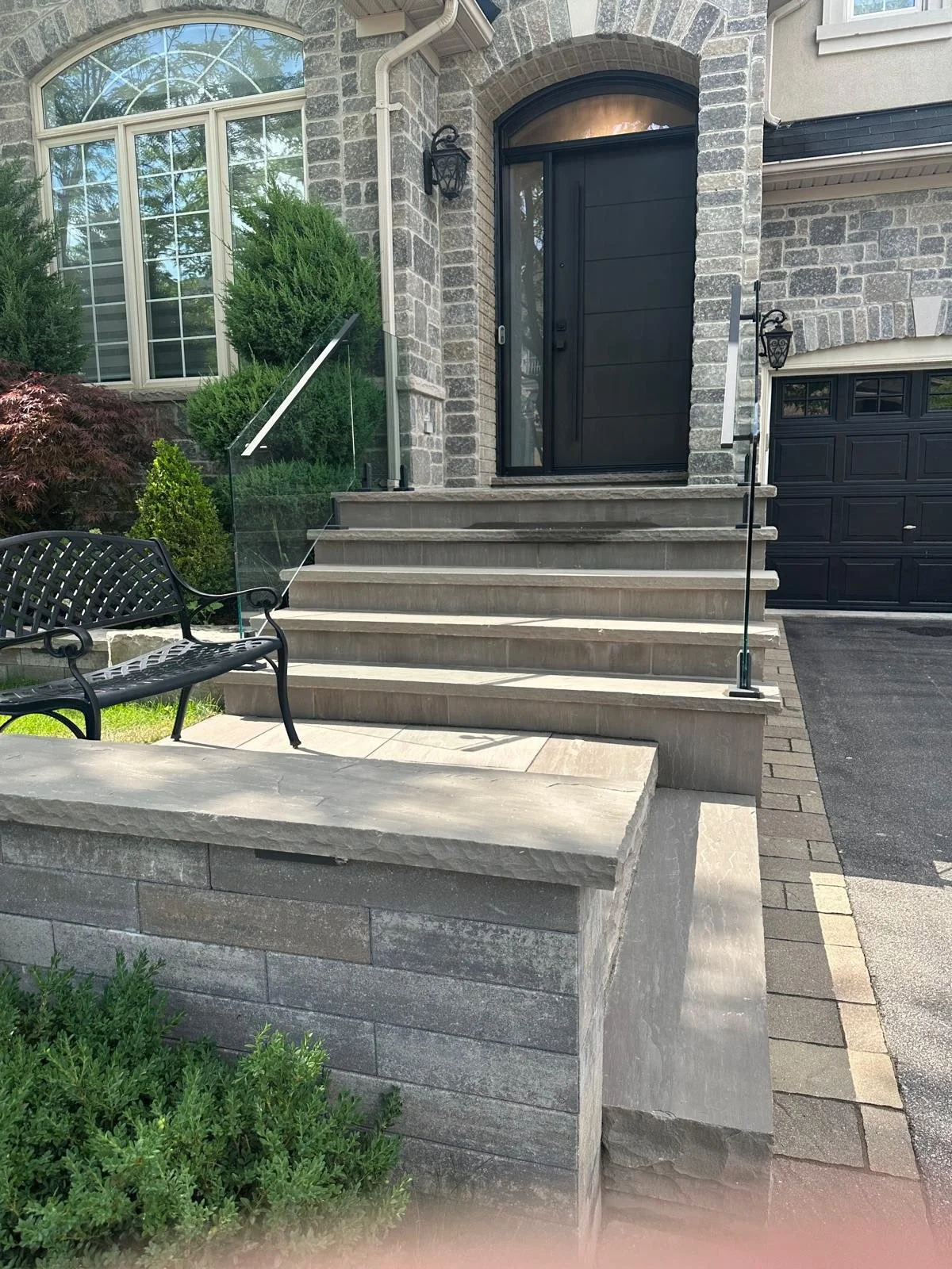 Front entrance of a house with a black door, stone steps, and a glass railing. Green bushes and a black metal bench are visible near the steps.