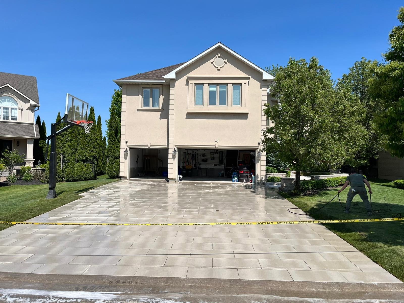 Newly washed and shiny concrete driveway in front of a beige two-story house with a garage, a basketball hoop on the left, and a man watering the grass on the right. Yellow caution tape marks off the area.