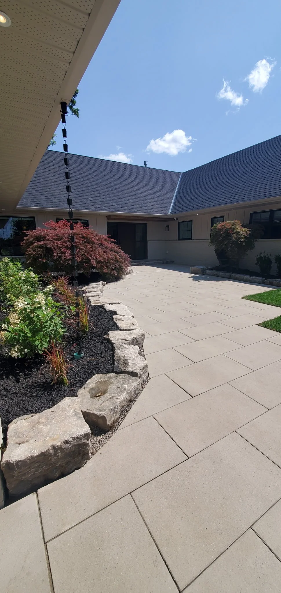 A courtyard with a paved walkway and garden beds, featuring rocks and plants, under a clear blue sky.