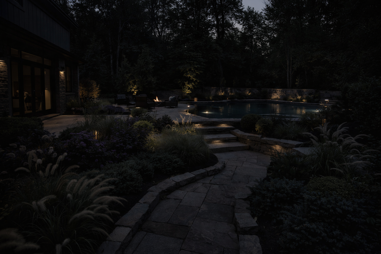 Nighttime view of a backyard with a lit swimming pool, surrounding patio furniture, and a garden with plants and flowers. The area is illuminated by landscape lighting and trees are visible in the background.