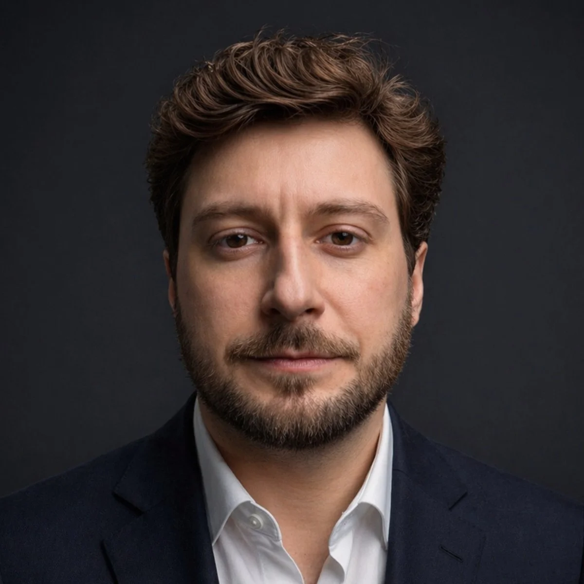 Close-up portrait of a young man with brown hair, a beard, wearing a white shirt and dark suit jacket, against a dark background. Rafael Leonardo Carlesso.