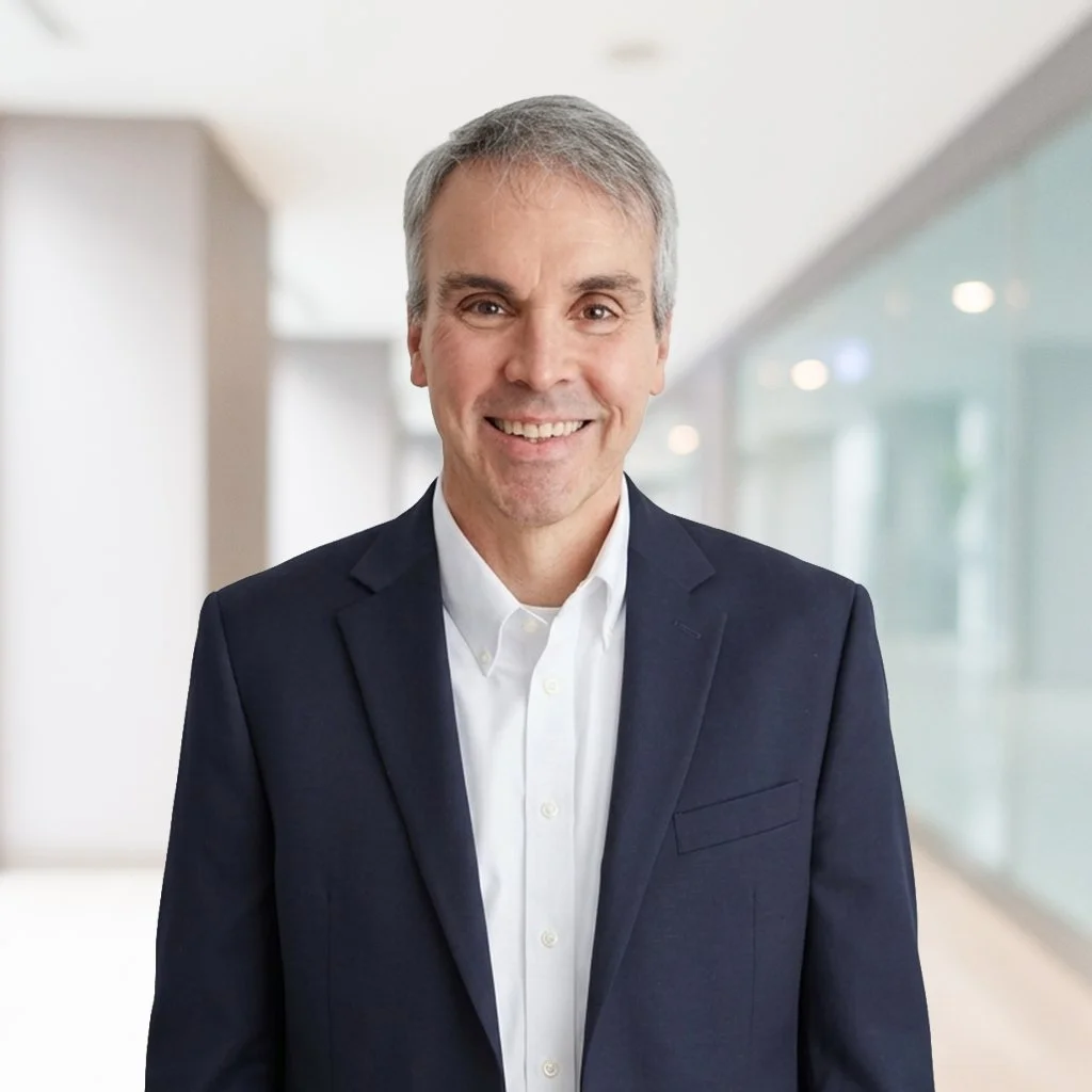 A smiling middle-aged man in a dark suit and white shirt standing in a modern, well-lit office or corridor with glass walls.
