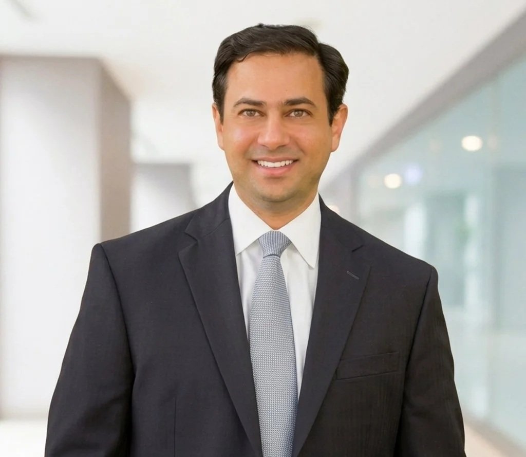 A smiling man in a dark suit, white shirt, and light gray tie standing in a modern office corridor.