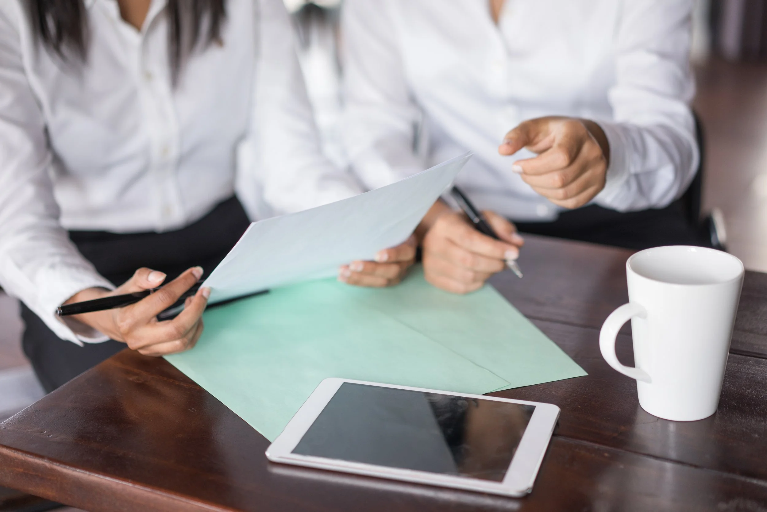 Deux personnes en chemise blanche discutent, une montre un document et l'autre pointe, sur une table en bois avec une tasse blanche et une tablette