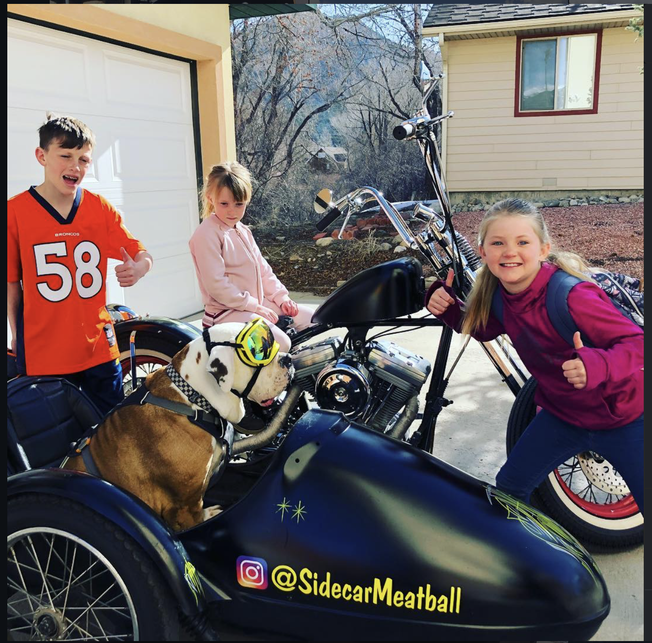 Three children and a dog posing with a motorcycle and sidecar. The boy on the left is wearing a Denver Broncos jersey and giving a thumbs up. The girl in the middle is sitting on the sidecar, and the girl on the right is leaning on the motorcycle, sm
