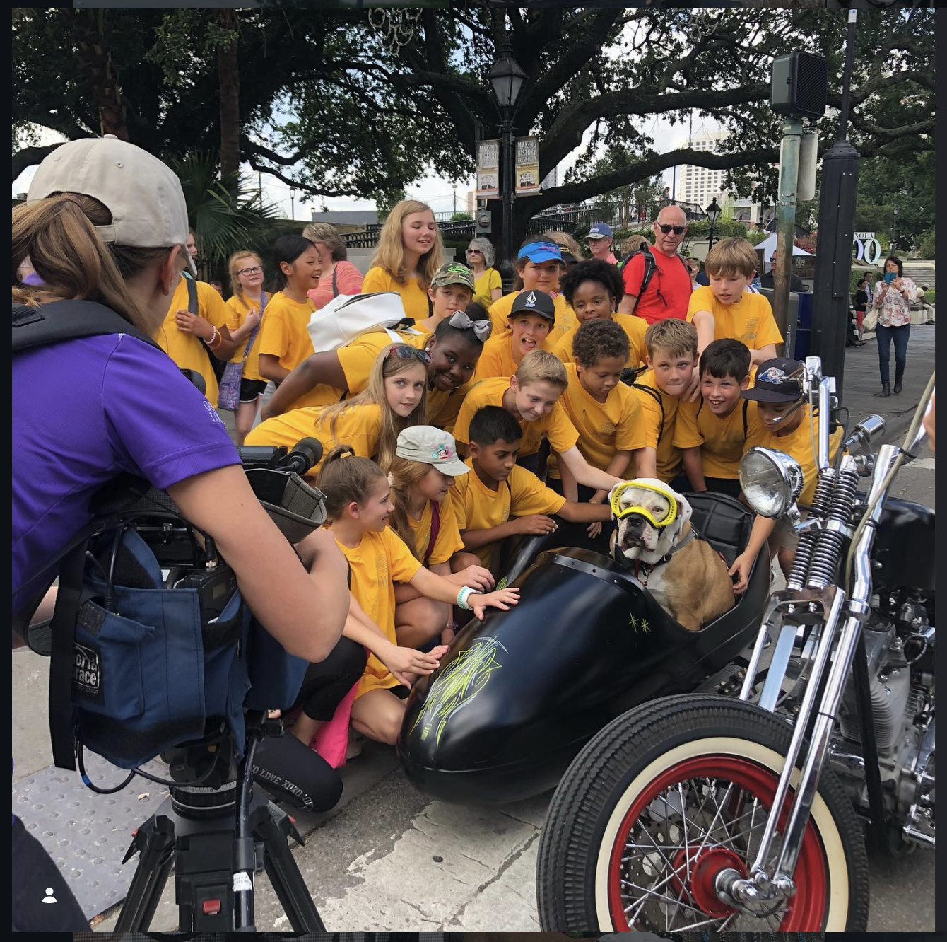 Group of children posing with a dog in a motorcycle sidecar, all wearing matching yellow shirts, under a large tree. A camera operator is filming the scene.