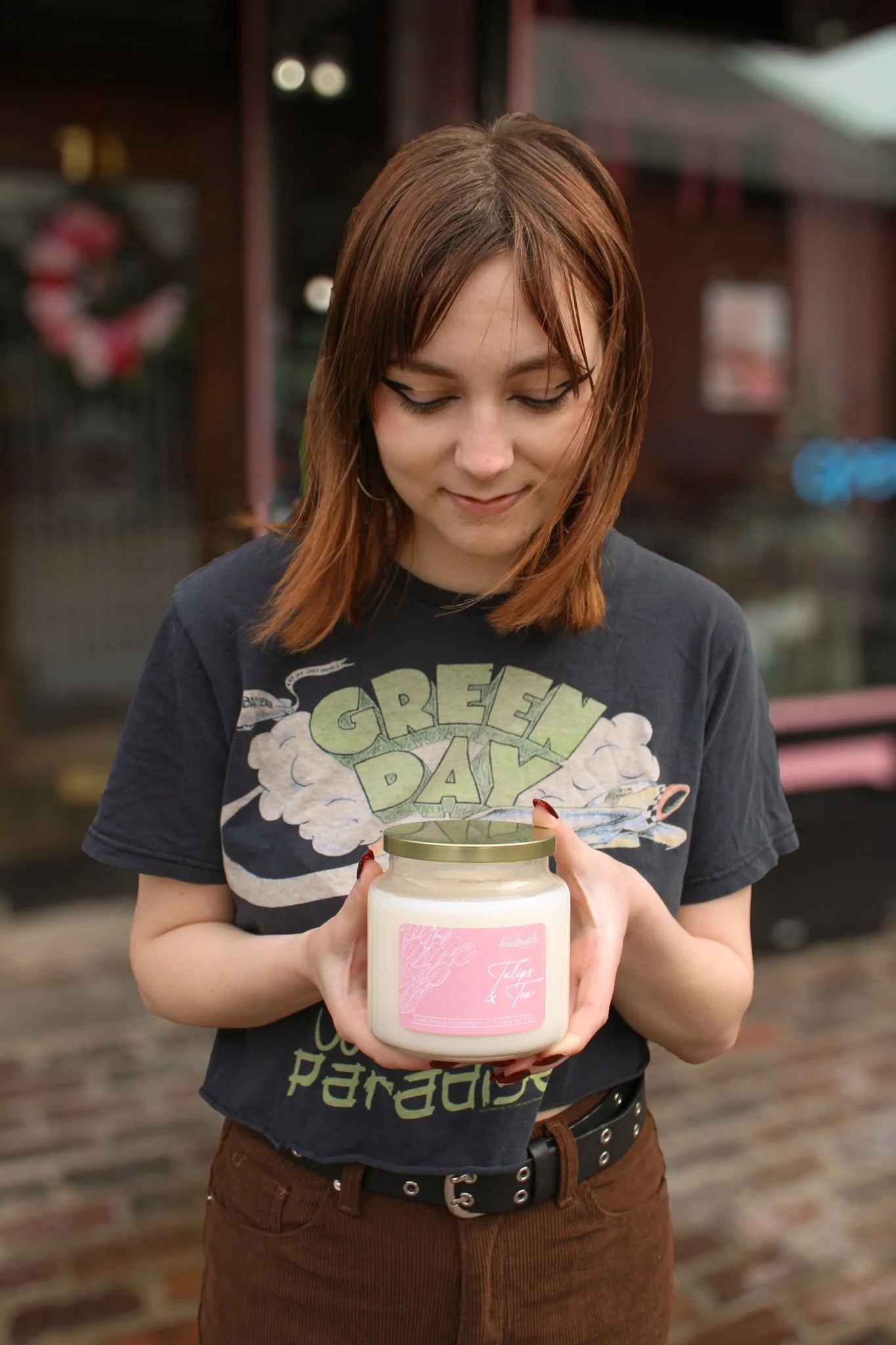 A young woman with shoulder-length brown hair and a black T-shirt with green and yellow lettering, holding a large pink-labeled candle jar, standing outdoors with a blurred background.