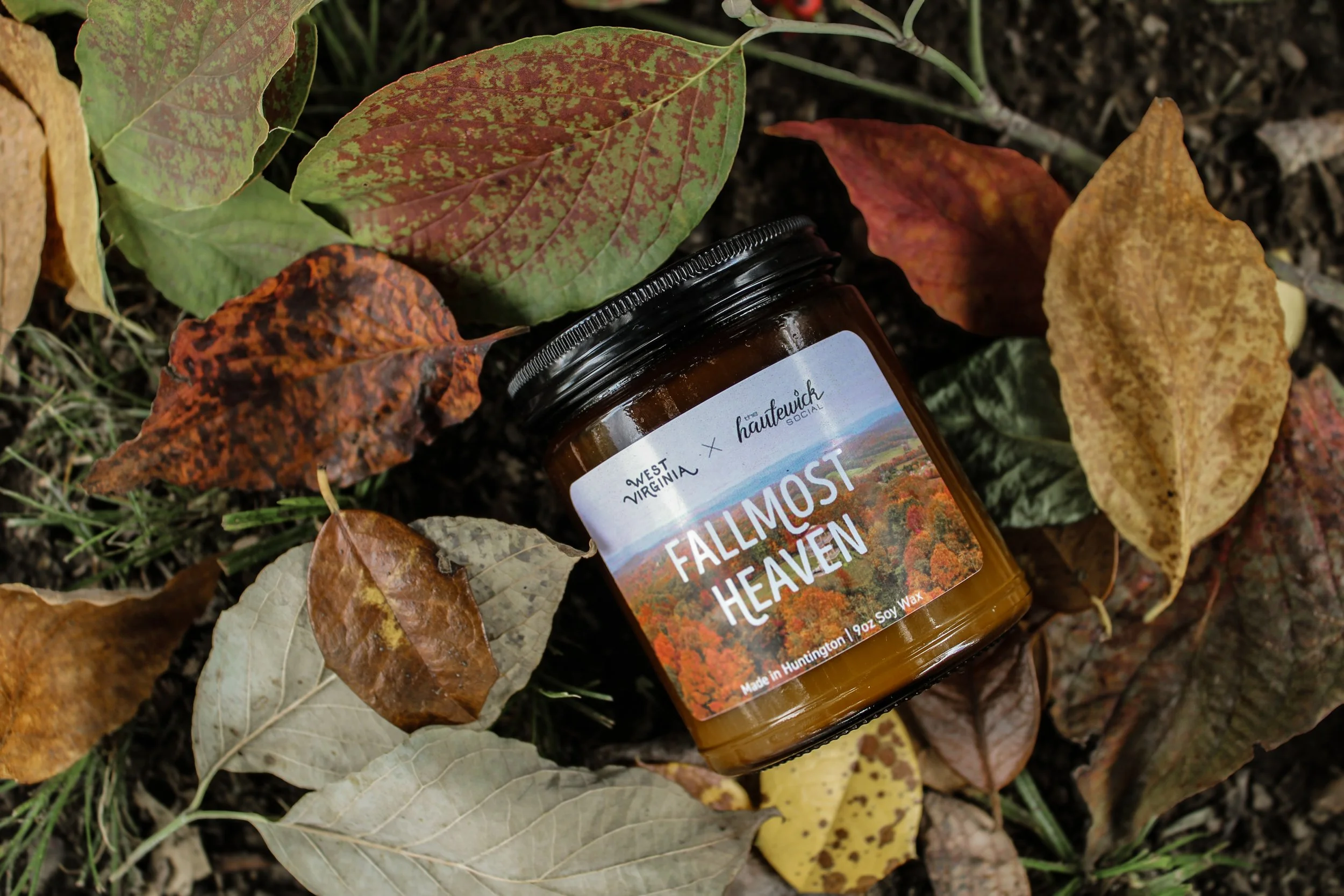A jar labeled 'Fallmost Heaven' lies on the ground amid fallen autumn leaves of various colors in a natural outdoor setting.