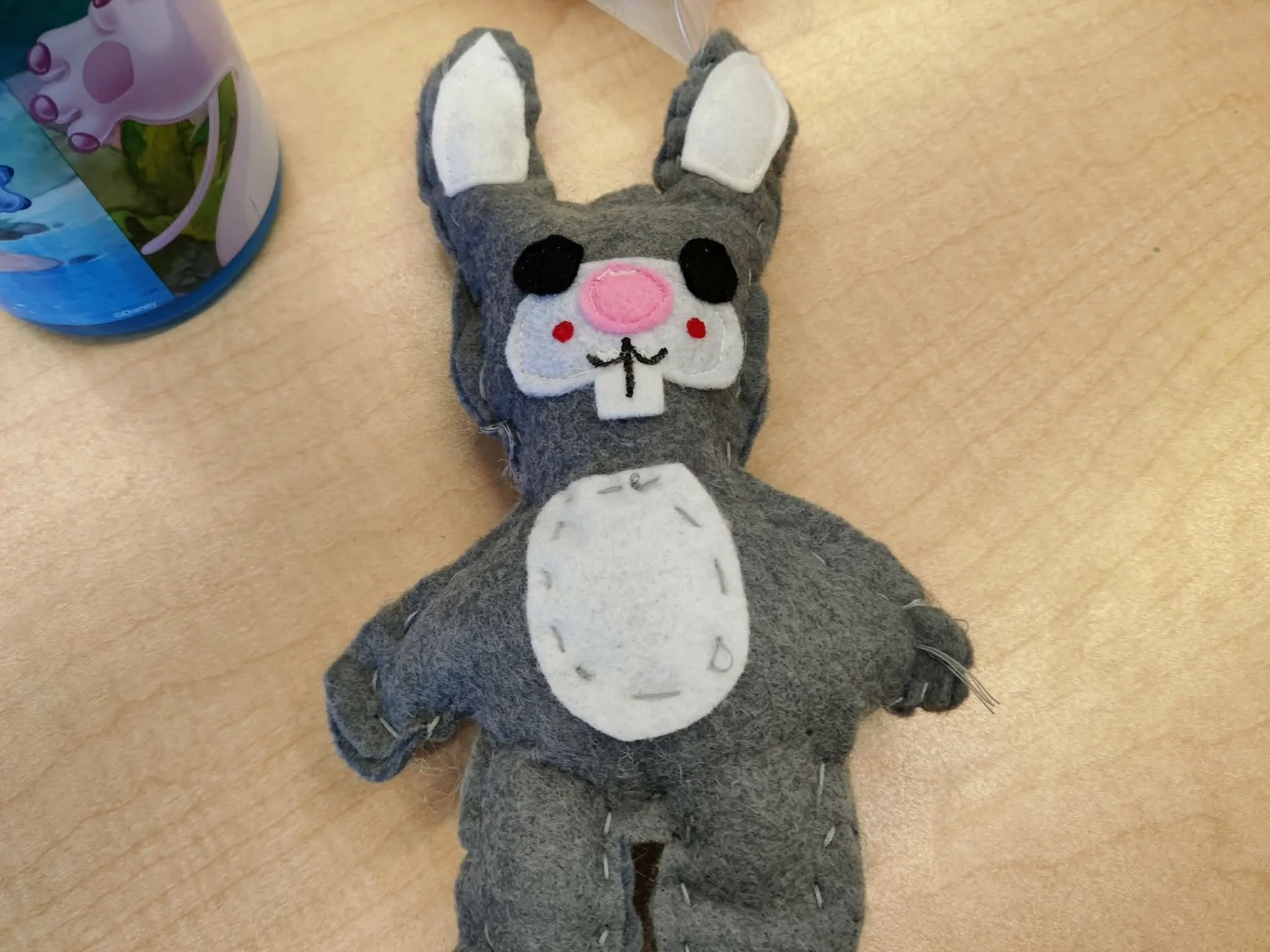 Une pantoufle en forme de chat gris avec un visage mignon, un nez rose, des pattes blanches, et des détails en feutrine noire, posée sur un sol en bois ou en linoléum beige.