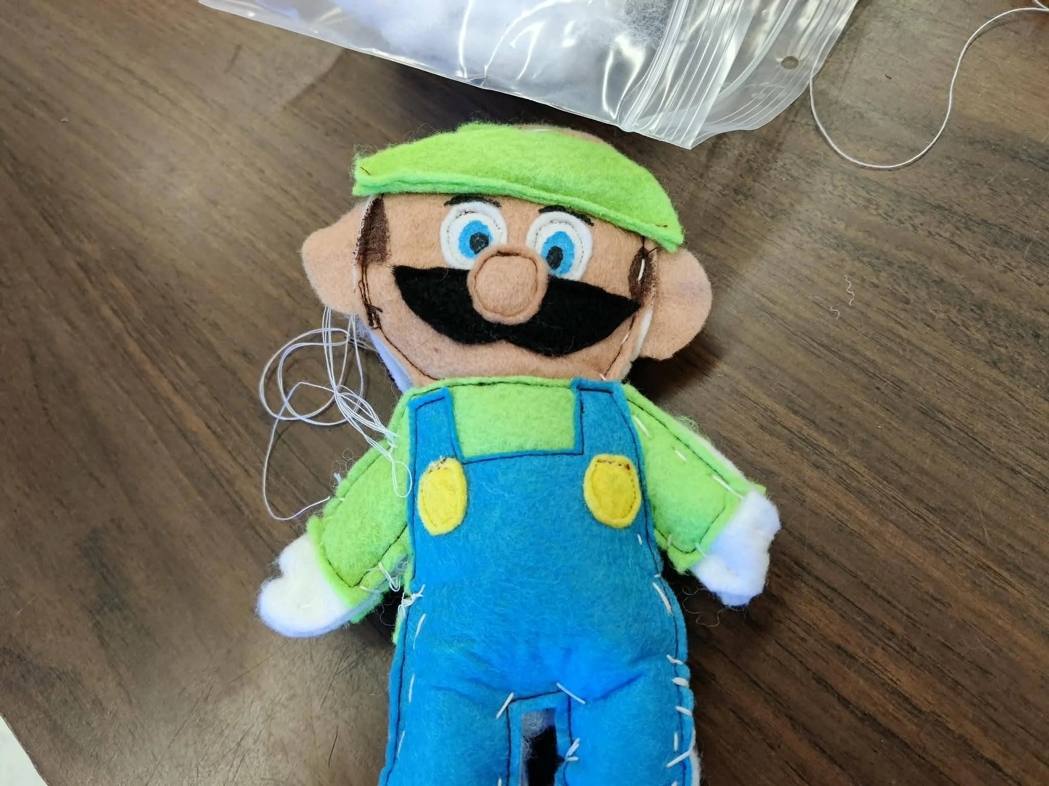 Marionnette en forme de personnage avec un chapeau vert, une moustache noire, des yeux bleus, et une salopette bleue, posée sur une surface en bois.