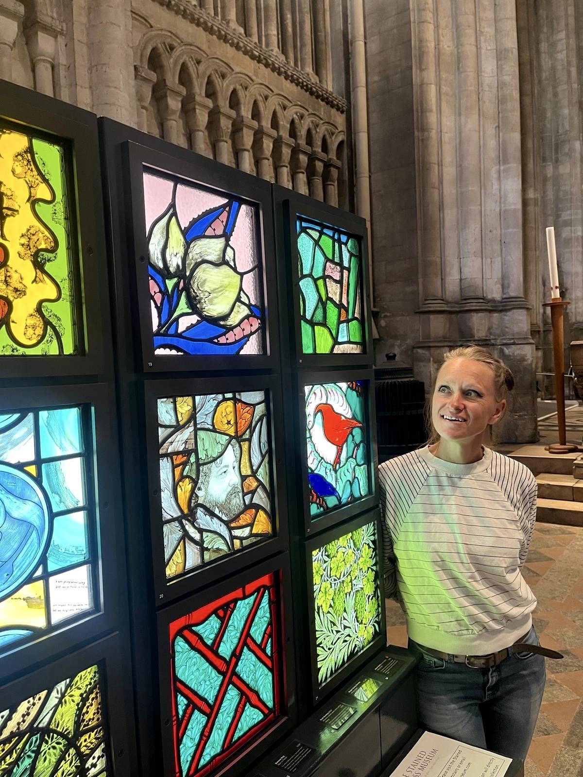 Zoe Henderson standing next to a display of colourful stained glass windows inside  Ely cathedral. Work displayed as part of the BSMP William Morris inspired painting on glass exhibition