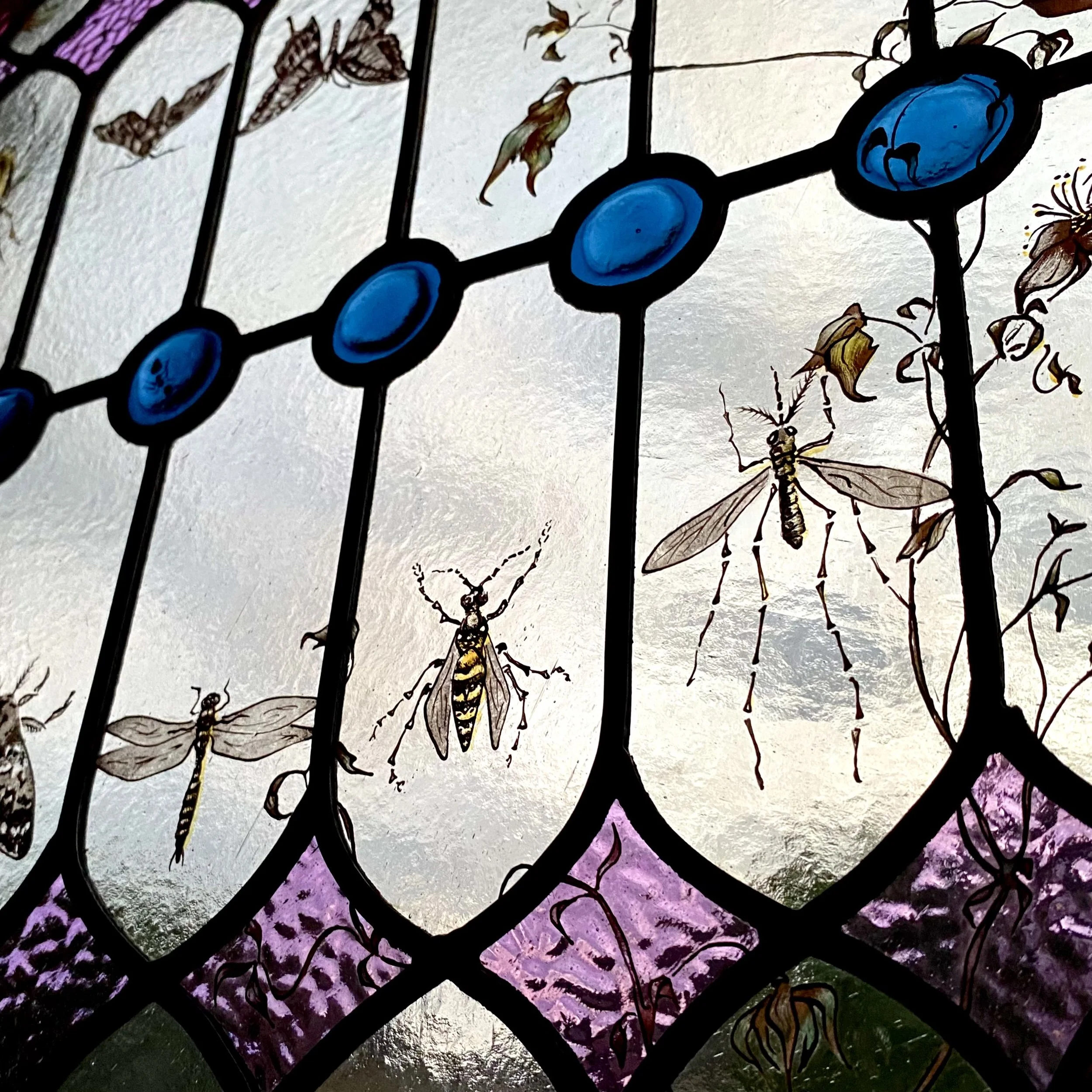 Close-up of a decorative stained glass window featuring painted insects, including dragonflies and bees, with a colorful background of purple, green, and light colors.