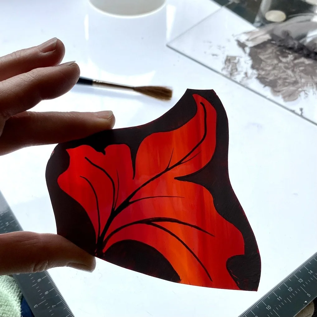 Painted decorative object resembling a red flower with black outlines, held by person's fingers, workspace with paintbrush, paper, and rulers in background.