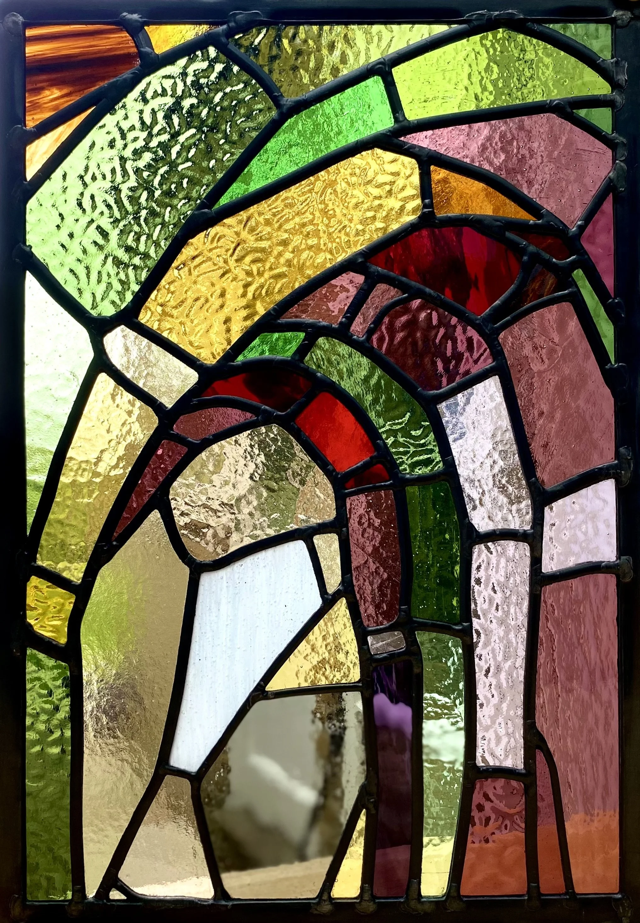 An abstract stained glass window featuring colourful, textured glass panes in various shapes and sizes.  The colors include green, yellow, red, orange, and purple, with some panes showing a textured surface.