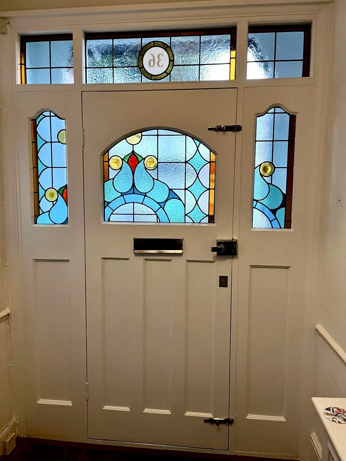 Best stained glass in london A white front door with decorative stained glass panels, featuring a circular design with the house number and some abstract patterns in blue, yellow, red, and green. Designed and built by Zoe Henderson South East London
